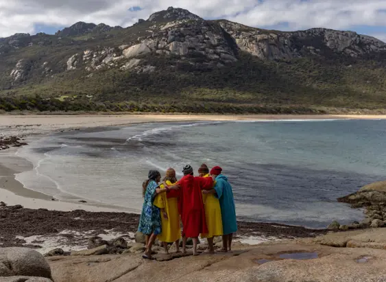 A group in colourful robes stand arm in arm on rocks overlooking a beach and mountains at FARAWAY on Flinders Island