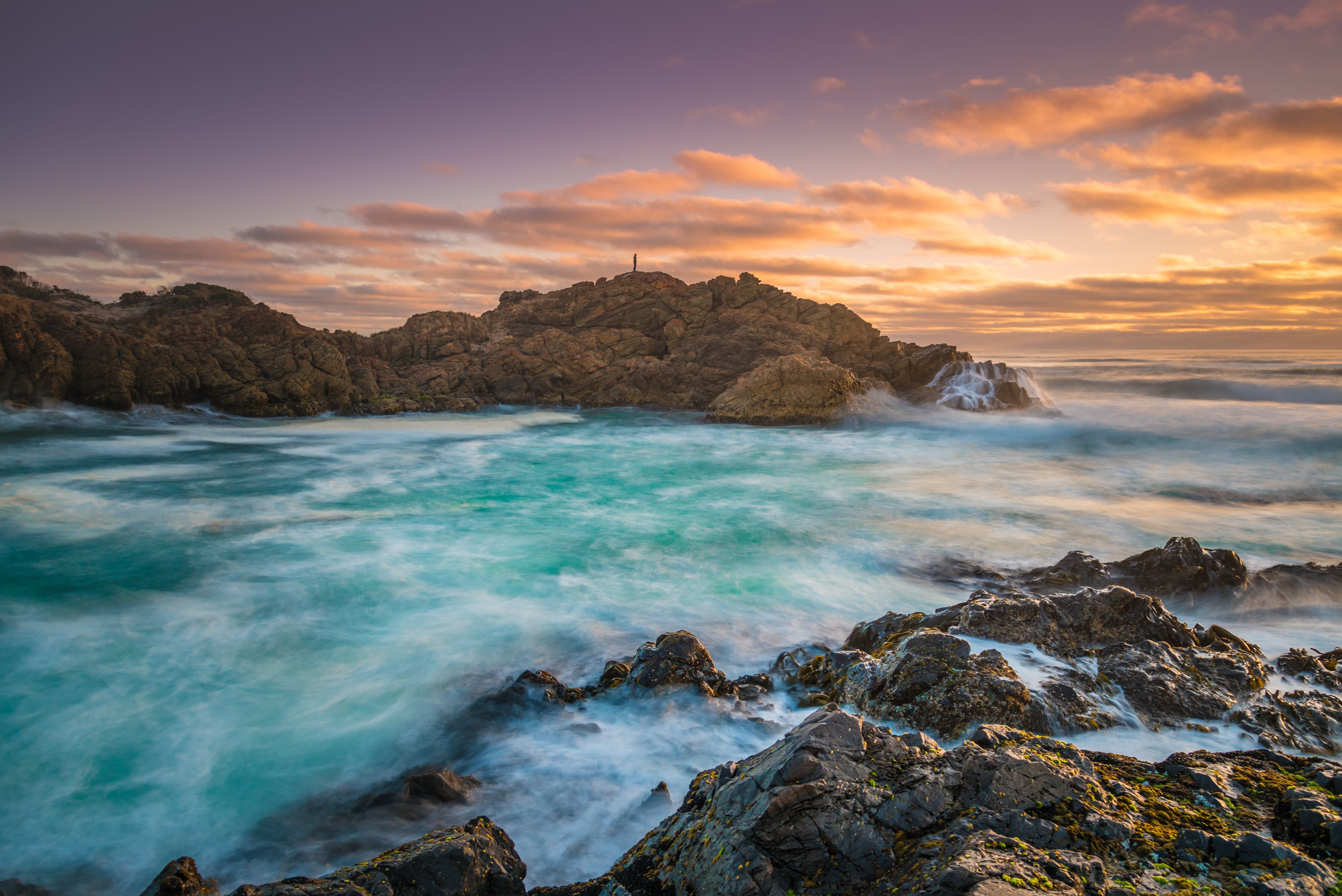 A person silhouetted on a rocky headland with waves breaking into a turquoise bay at Trial Harbour, Tasmania's West Coast