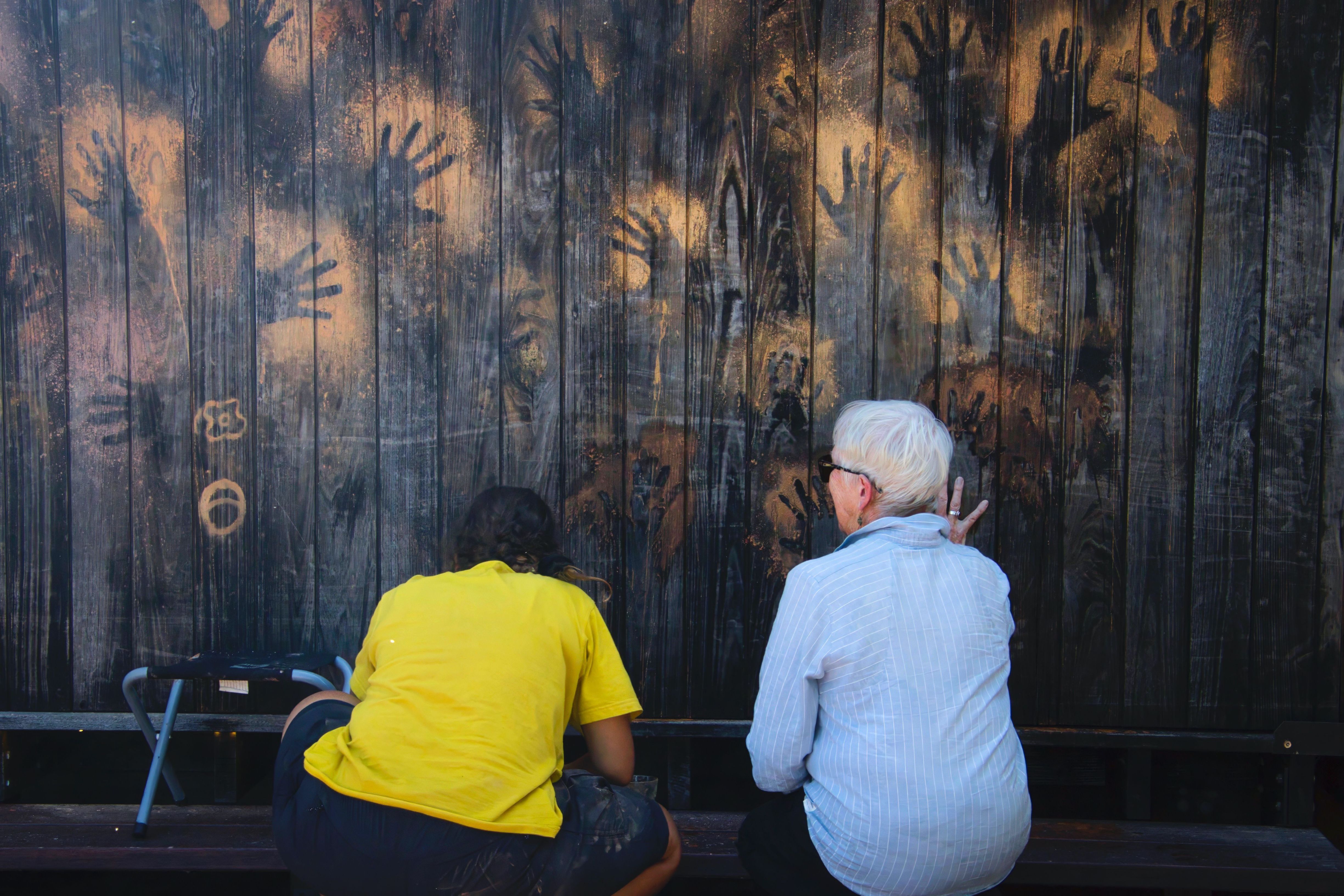 Two people sit with their backs to camera looking at ochre handprints on a dark wooden wall on the wukalina / Mount William walk