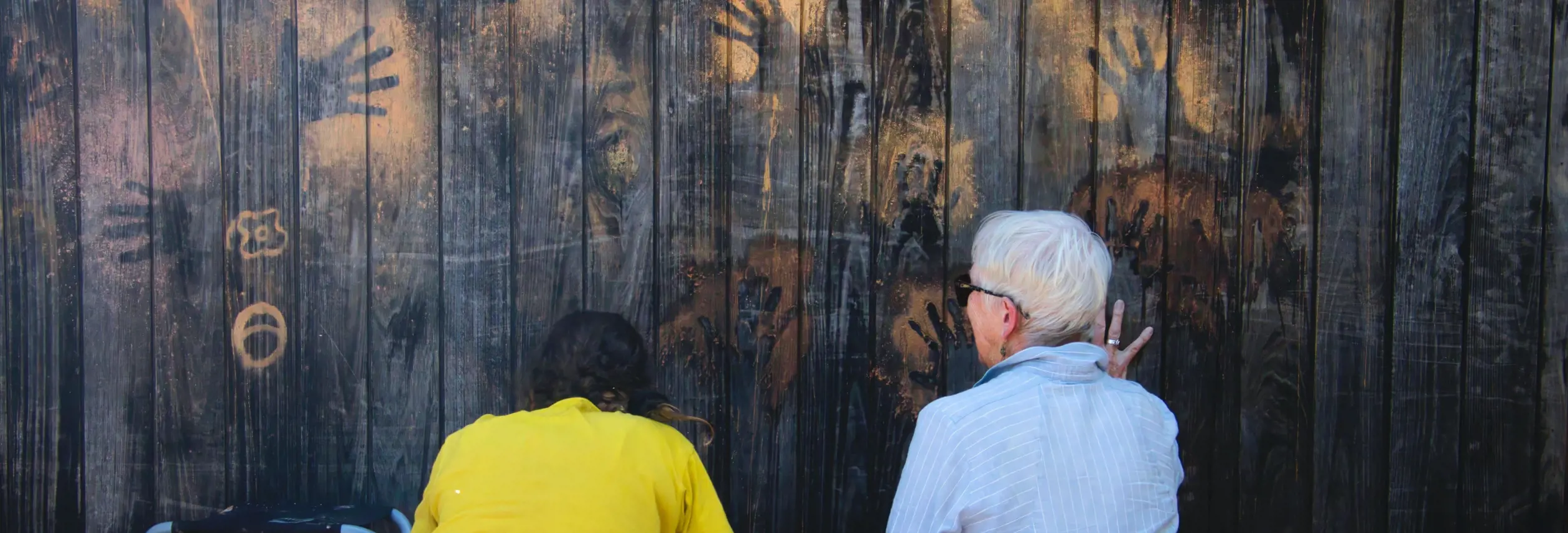 Two people sit with their backs to camera looking at ochre handprints on a dark wooden wall on the wukalina / Mount William walk