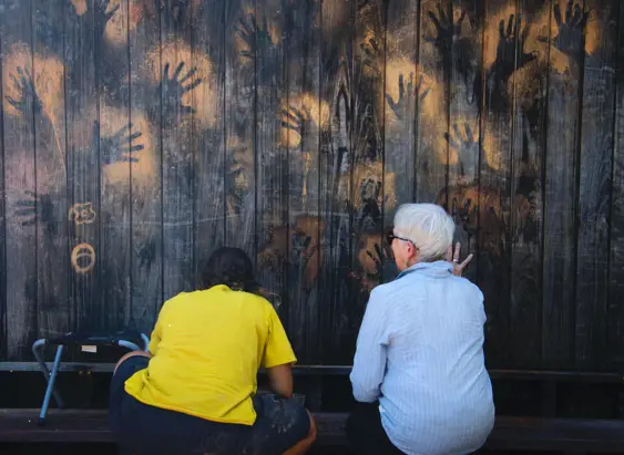 Two people sit with their backs to camera looking at ochre handprints on a dark wooden wall on the wukalina / Mount William walk