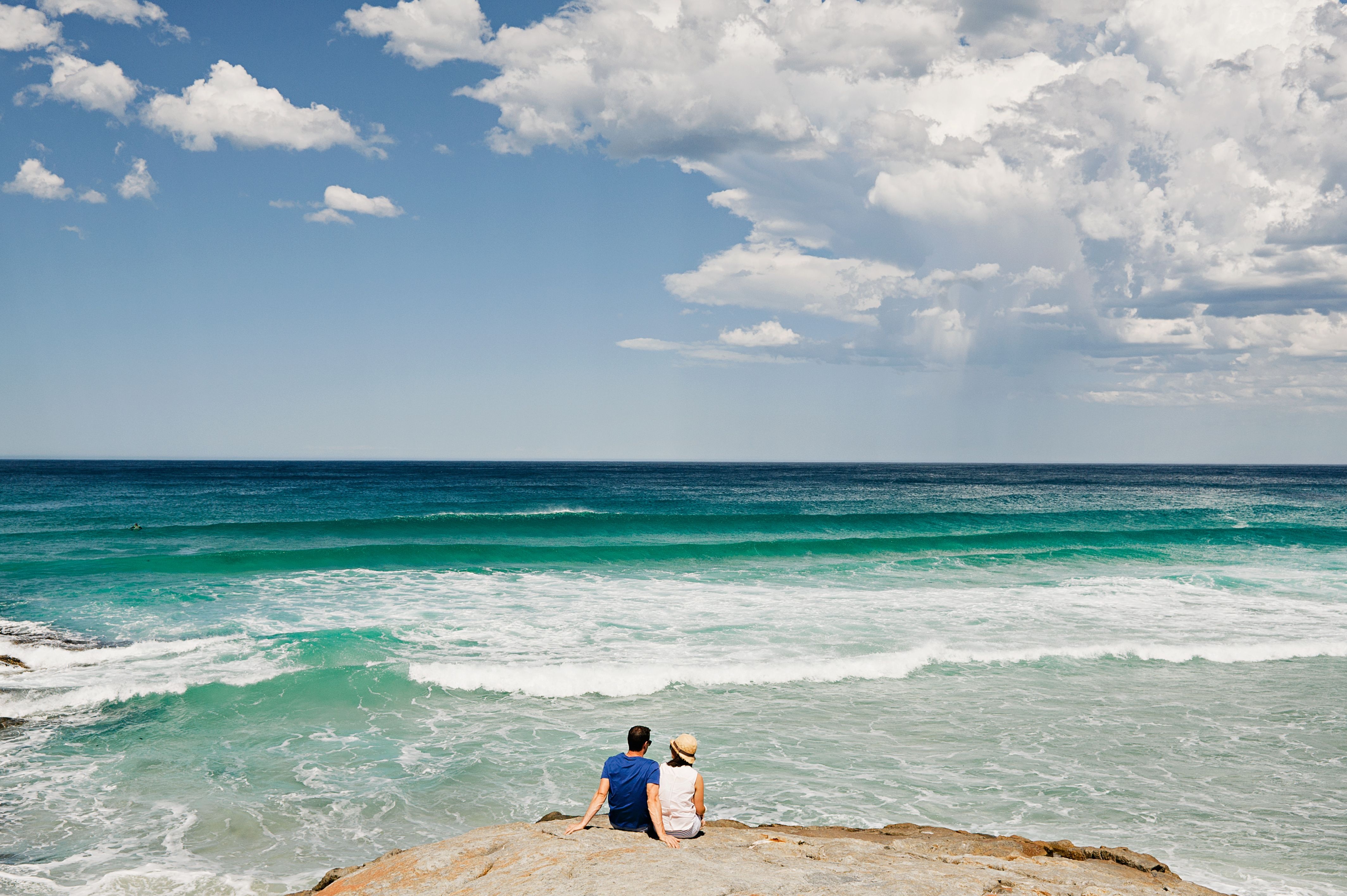 Two people sitting on coastal rocks watching turquoise waves at Little Beach, Tasmania's East Coast