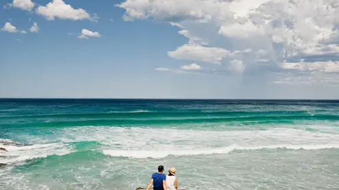 Two people sitting on coastal rocks watching turquoise waves at Little Beach, Tasmania's East Coast