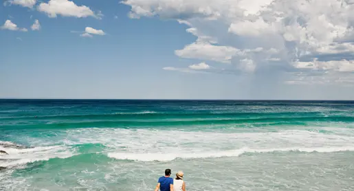 Two people sitting on coastal rocks watching turquoise waves at Little Beach, Tasmania's East Coast
