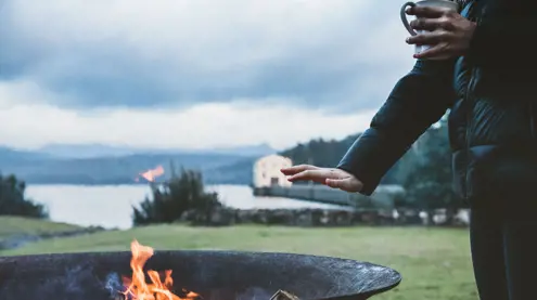 A guest with a mug warming hands by a firepit, Pumphouse Point visible in the background, Lake St Clair, Tasmania
