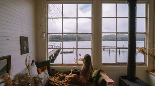 A guest with a mug looking through windows toward the water and jetty at Captain's Rest, Strahan on Tasmania's West Coast