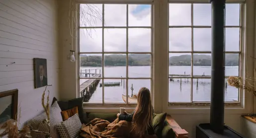 A guest with a mug looking through windows toward the water and jetty at Captain's Rest, Strahan on Tasmania's West Coast