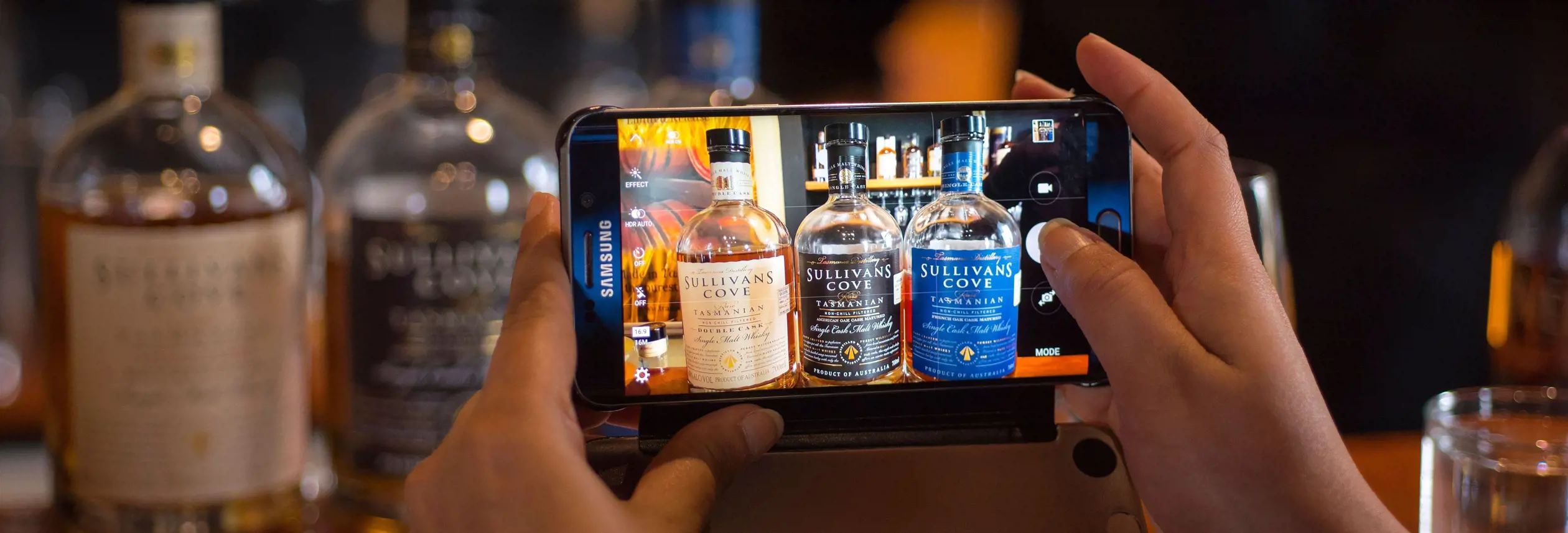 Hands holding a phone photographing Tasmanian whisky bottles including Sullivans Cove, Hobart, Tasmania