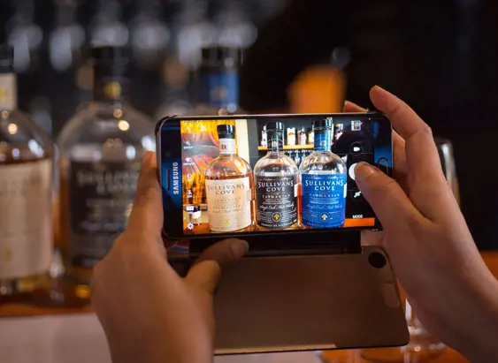 Hands holding a phone photographing Tasmanian whisky bottles including Sullivans Cove, Hobart, Tasmania