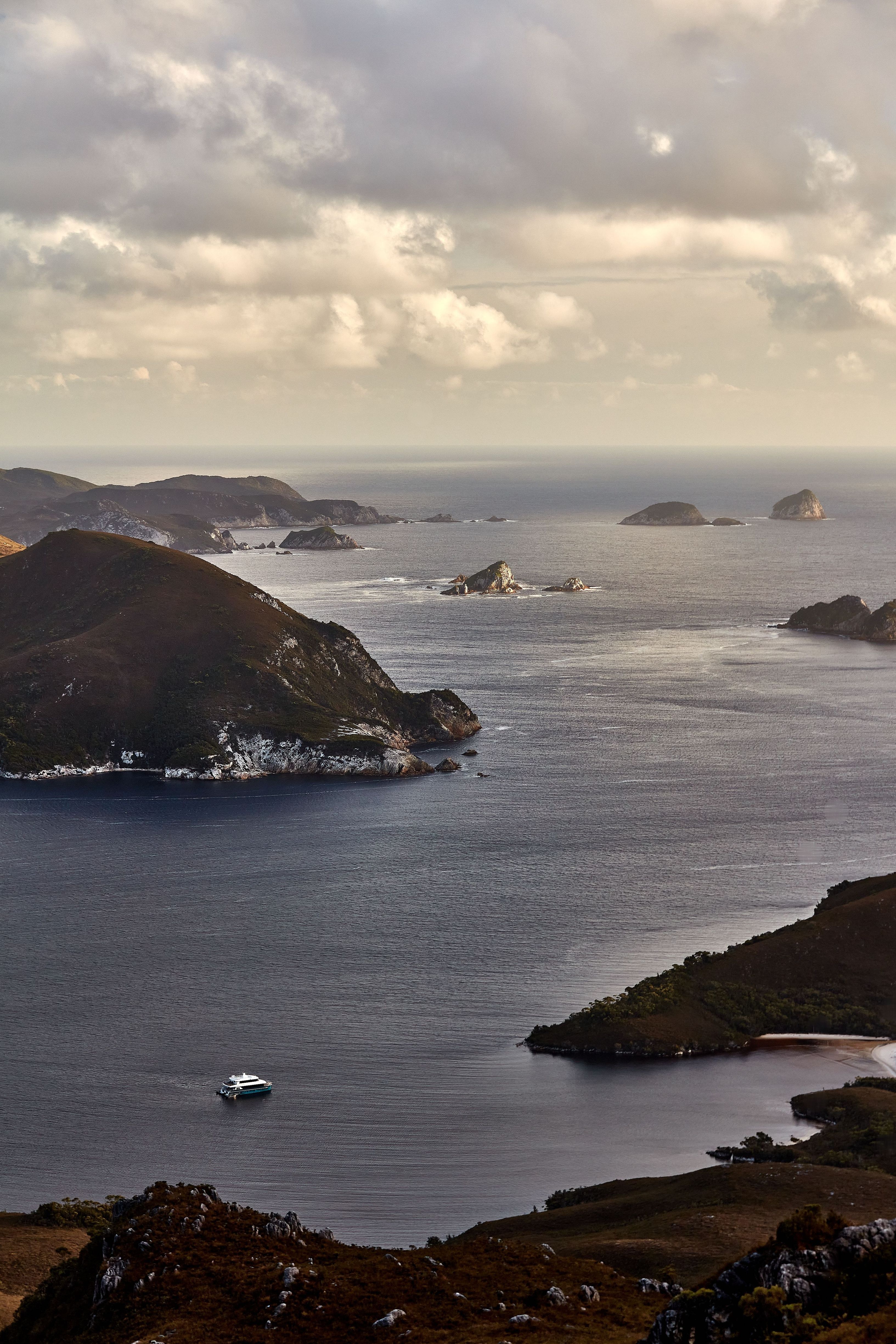 Aerial view of an On Board expedition vessel in Bathurst Harbour with rocky islets and headlands, Southwest National Park, Tasmania