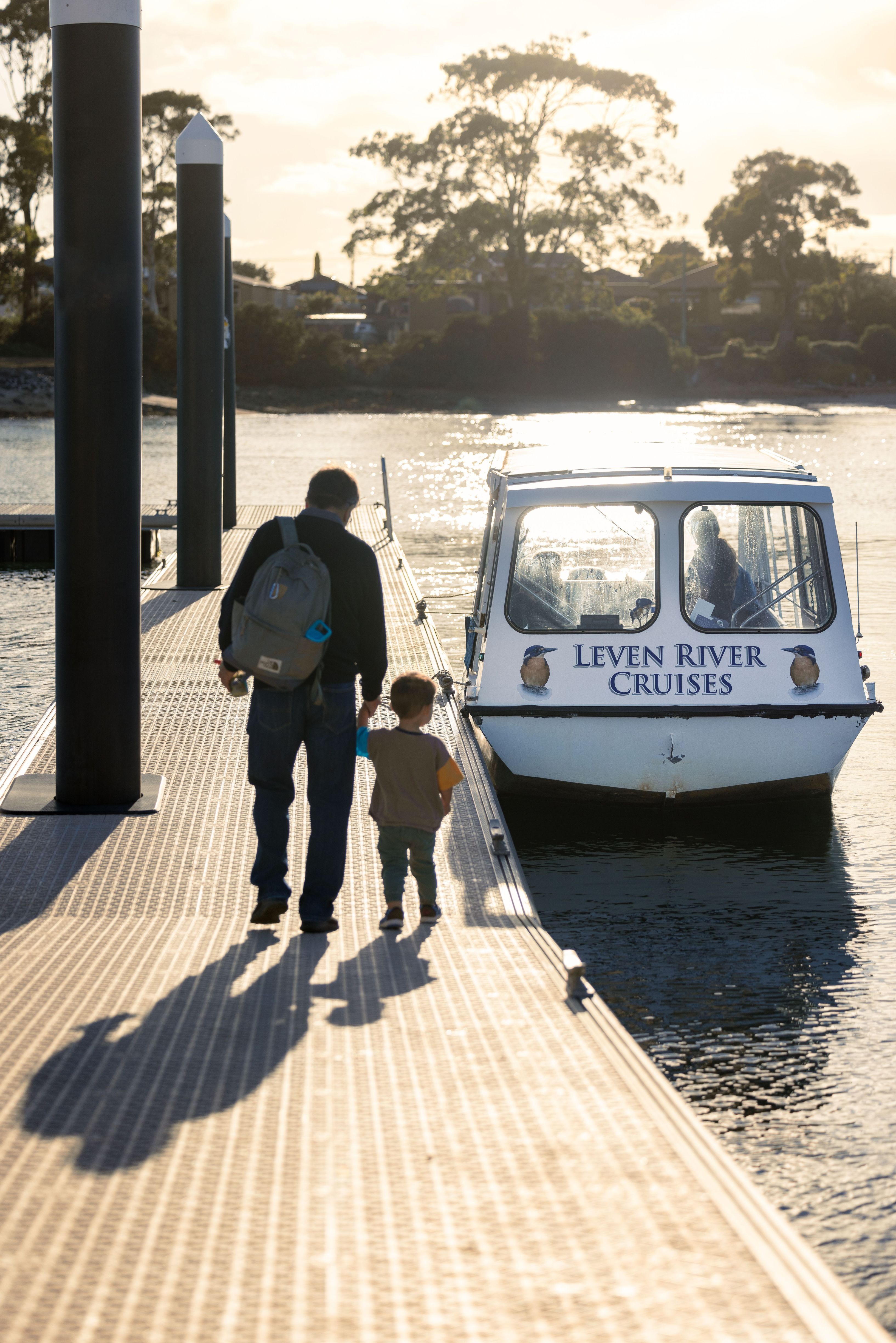 An adult and child walking along the jetty towards Leven River Cruises boat at golden hour, Ulverstone, Tasmania