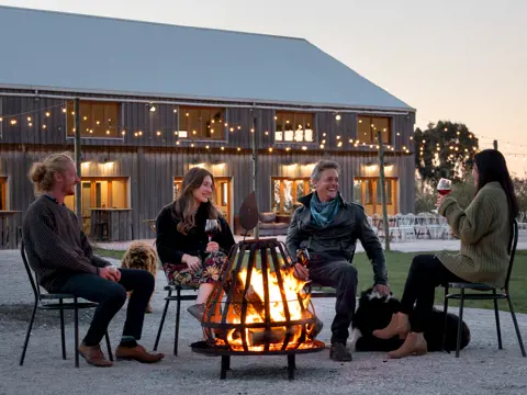 Guests gathered around a fire pit with wine and dogs outside the barn at Mount Gnomon Farm at dusk, Penguin, Tasmania