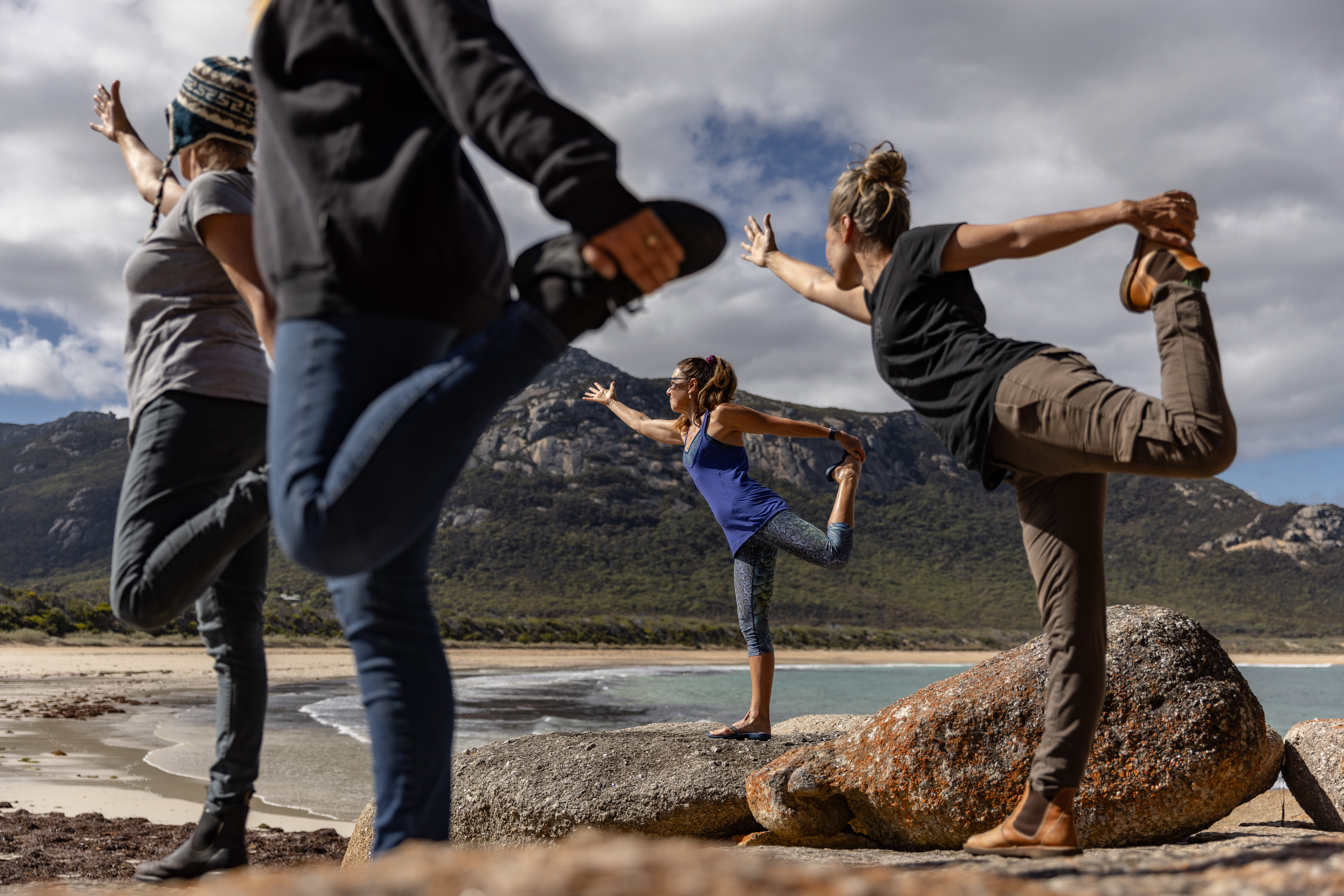 Four people practising yoga on rocks in Lady Barron with a beach and mountains behind, FARAWAY on Flinders Island, Tasmania