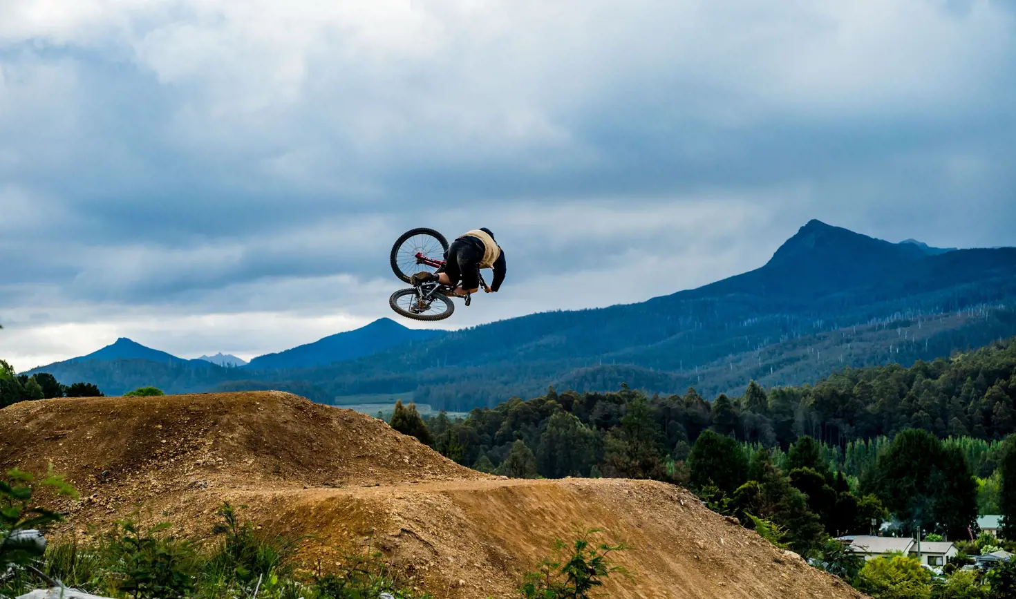 A mountain biker mid-jump on a dirt track with mountains behind at Maydena Bike Park, Derwent Valley, Tasmania