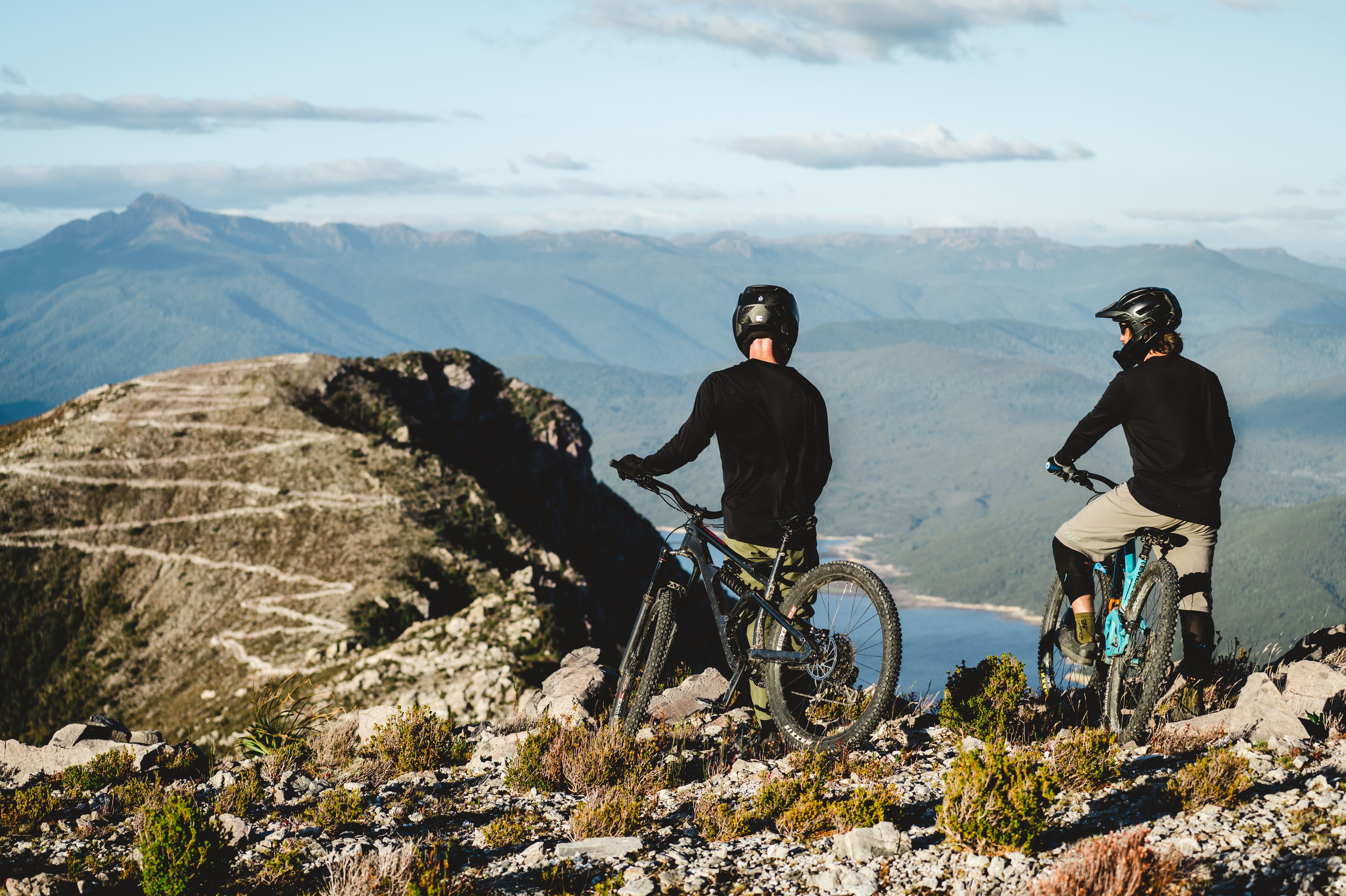Two mountain bikers on a rocky outcrop overlooking mountains and water at Mount Owen, Tasmania's West Coast