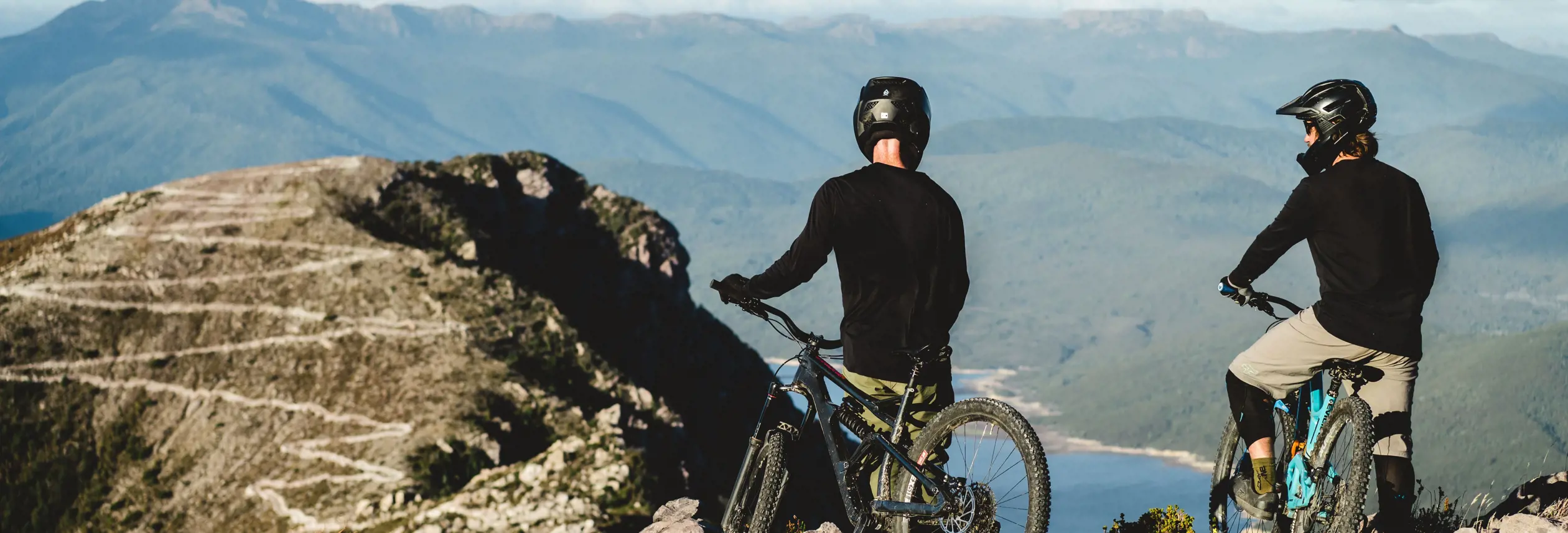 Two mountain bikers on a rocky outcrop overlooking mountains and water at Mount Owen, Tasmania's West Coast