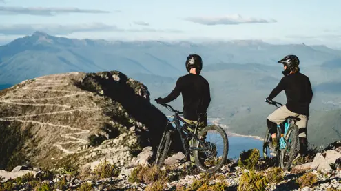 Two mountain bikers on a rocky outcrop overlooking mountains and water at Mount Owen, Tasmania's West Coast