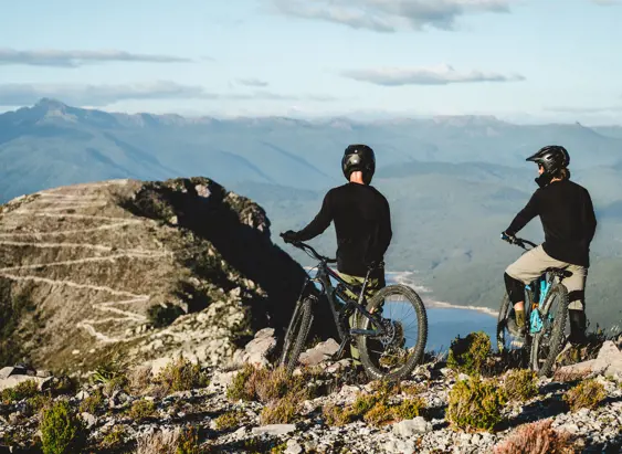 Two mountain bikers on a rocky outcrop overlooking mountains and water at Mount Owen, Tasmania's West Coast