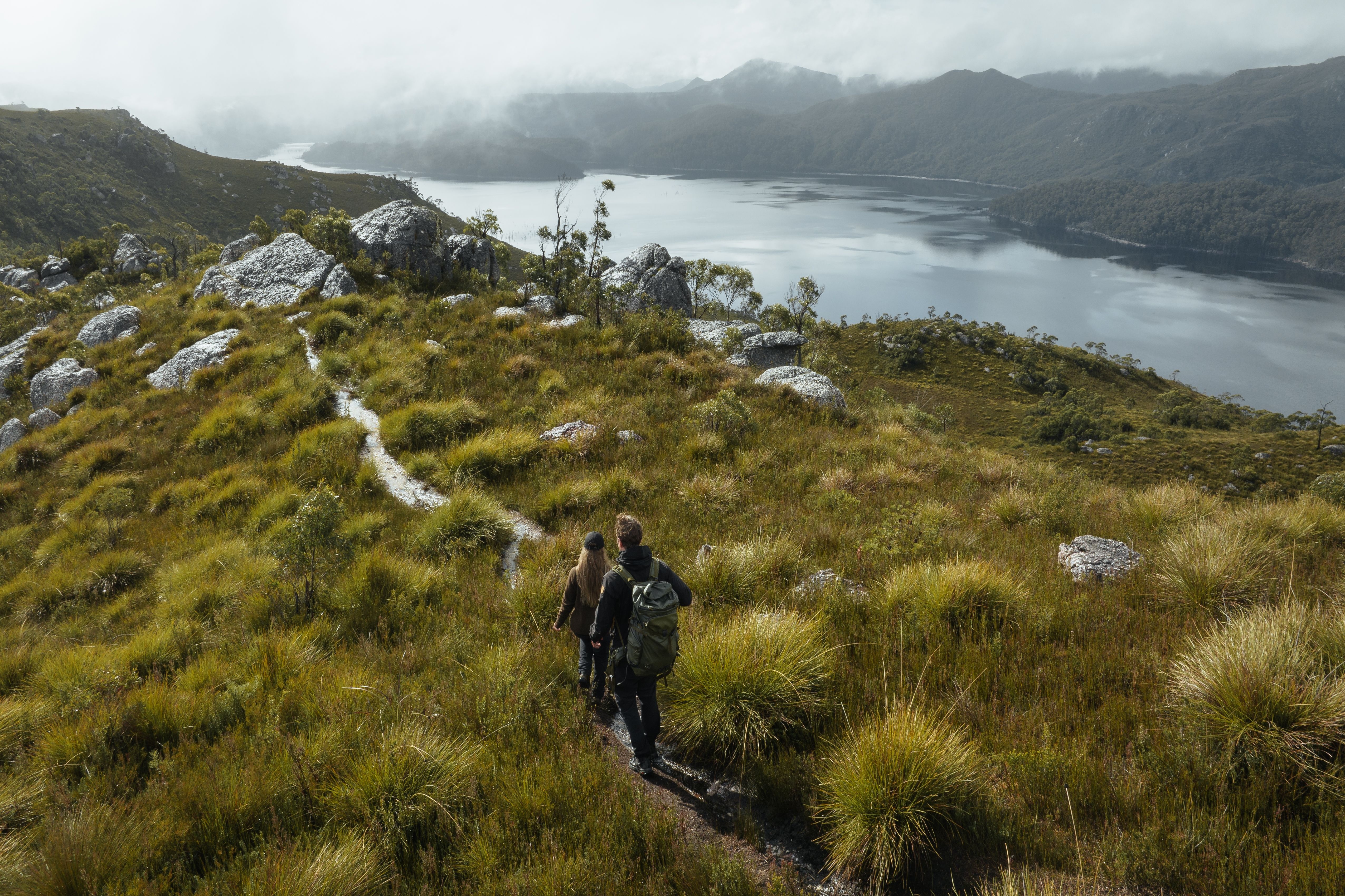 Two walkers on a track through grassland with a body of water and misty mountains beyond at Mount Farrell, West Coast Range, Tasmania