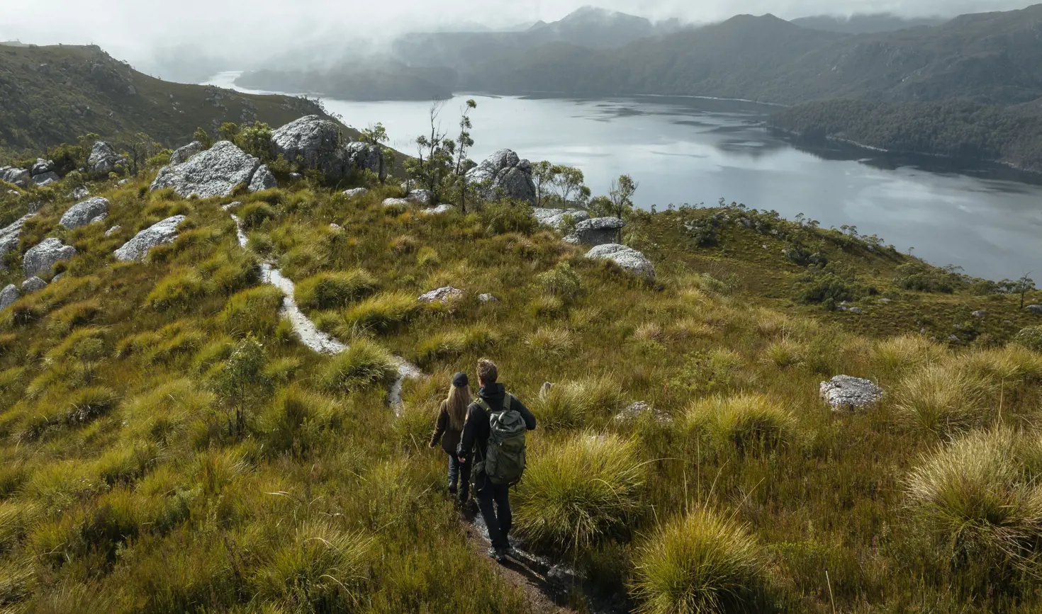 Two walkers on a track through grassland with a body of water and misty mountains beyond at Mount Farrell, West Coast Range, Tasmania