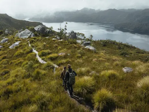 Two walkers on a track through grassland with a body of water and misty mountains beyond at Mount Farrell, West Coast Range, Tasmania