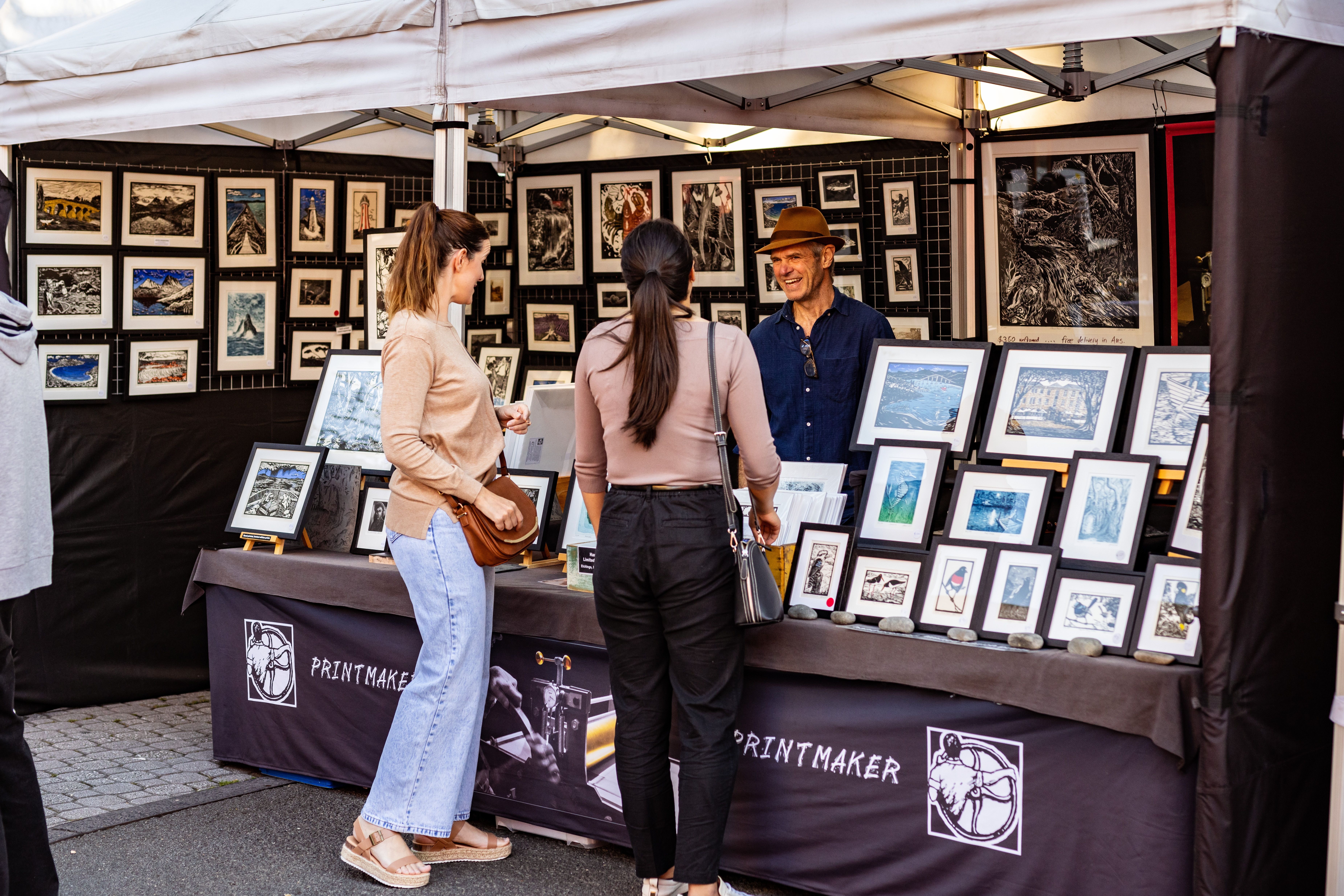 Two customers browsing framed artworks at the Printmaker stall at Salamanca Market, Hobart, Tasmania
