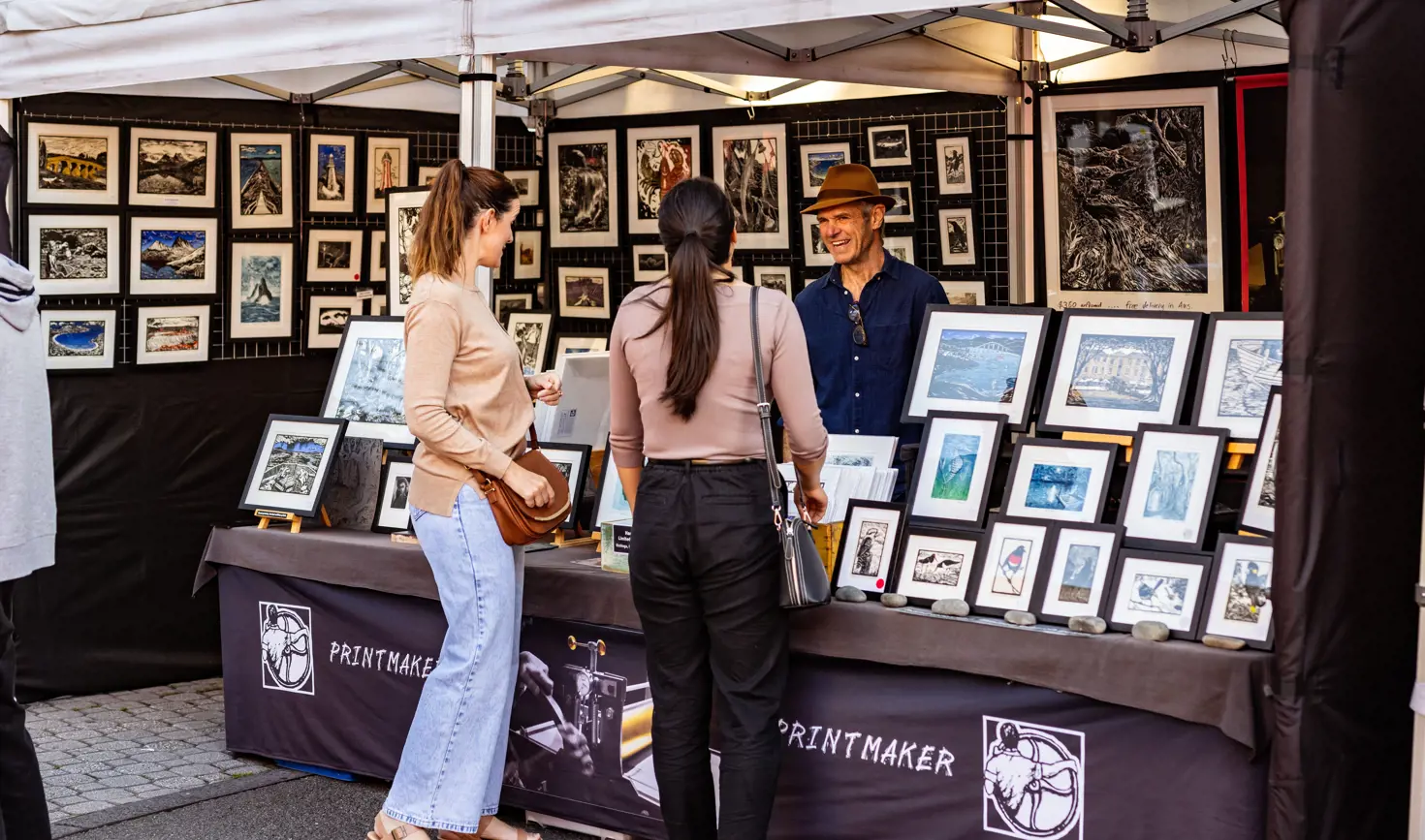 Two customers browsing framed artworks at the Printmaker stall at Salamanca Market, Hobart, Tasmania