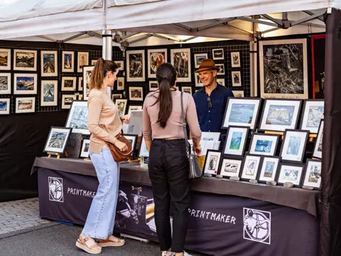 Two customers browsing framed artworks at the Printmaker stall at Salamanca Market, Hobart, Tasmania