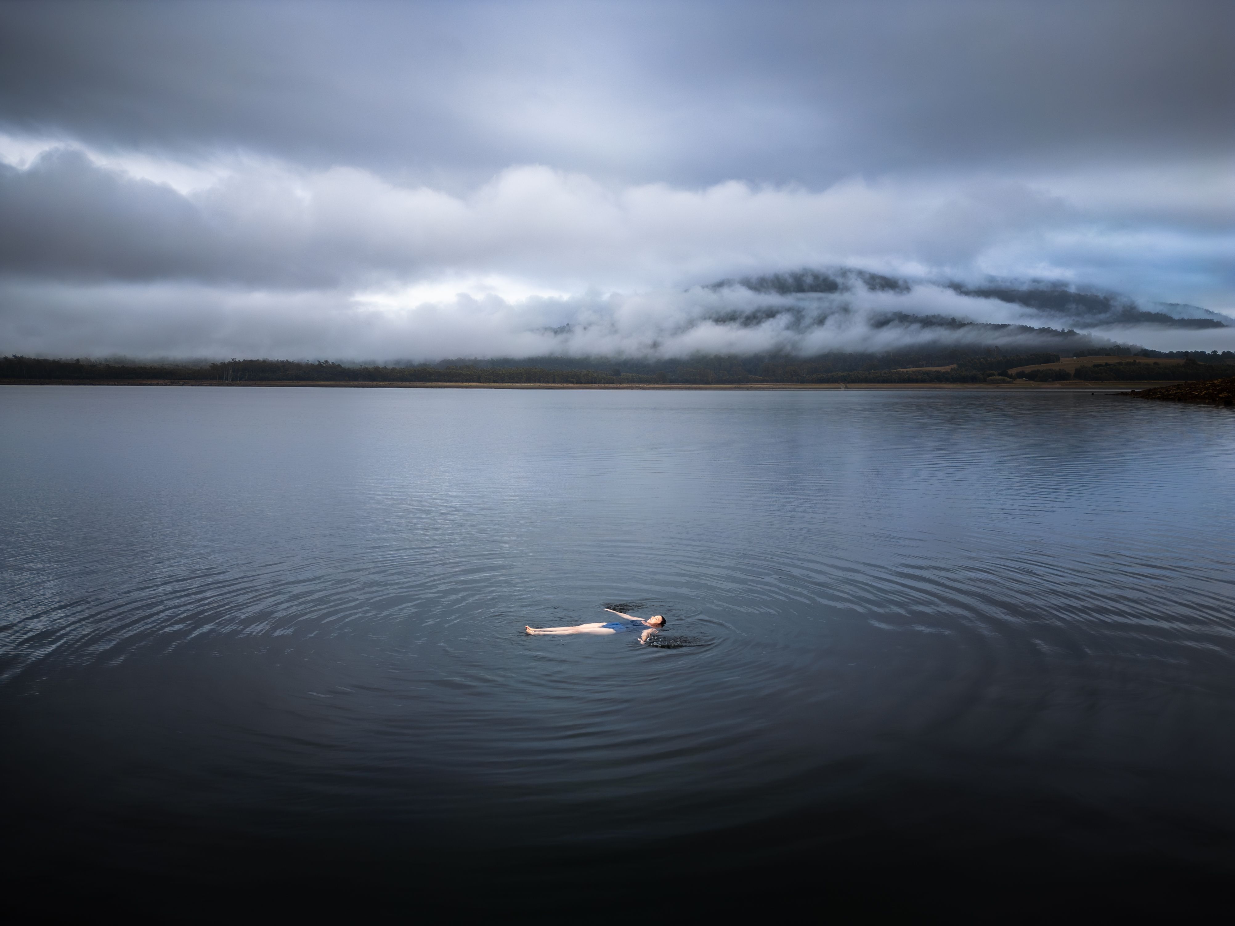 A swimmer floating on their back in misty calm water with cloud covered hills beyond, near Cedar Cottage Meander, Tasmania