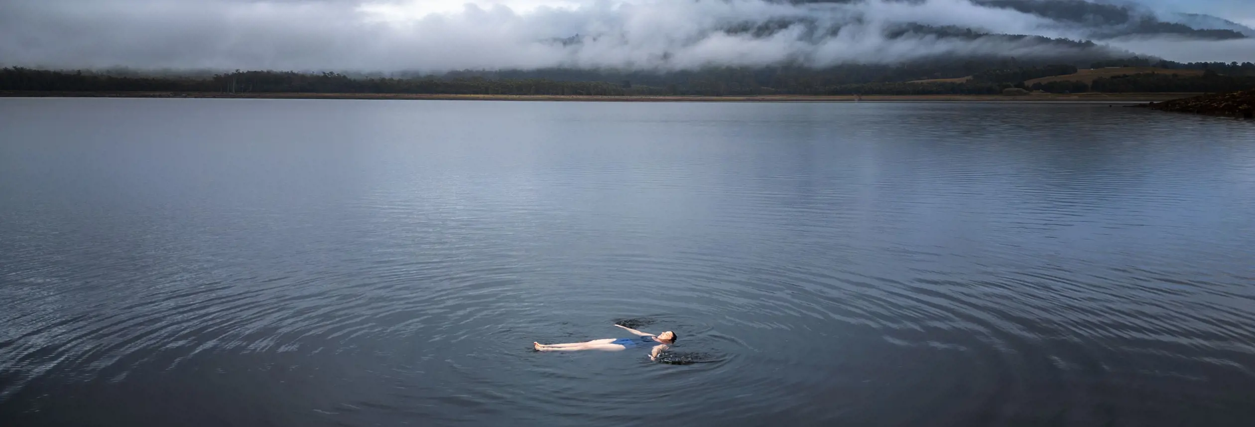 A swimmer floating on their back in misty calm water with cloud covered hills beyond, near Cedar Cottage Meander, Tasmania