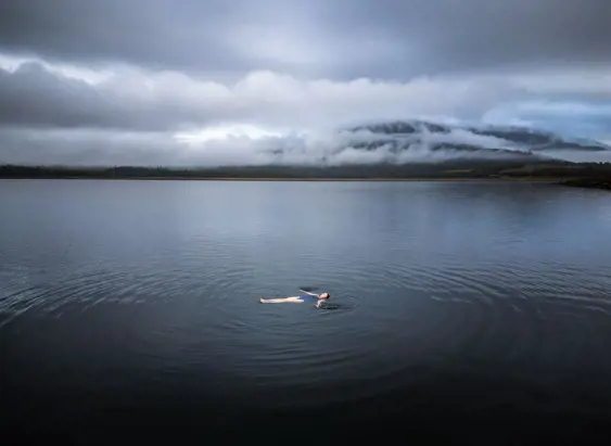 A swimmer floating on their back in misty calm water with cloud covered hills beyond, near Cedar Cottage Meander, Tasmania