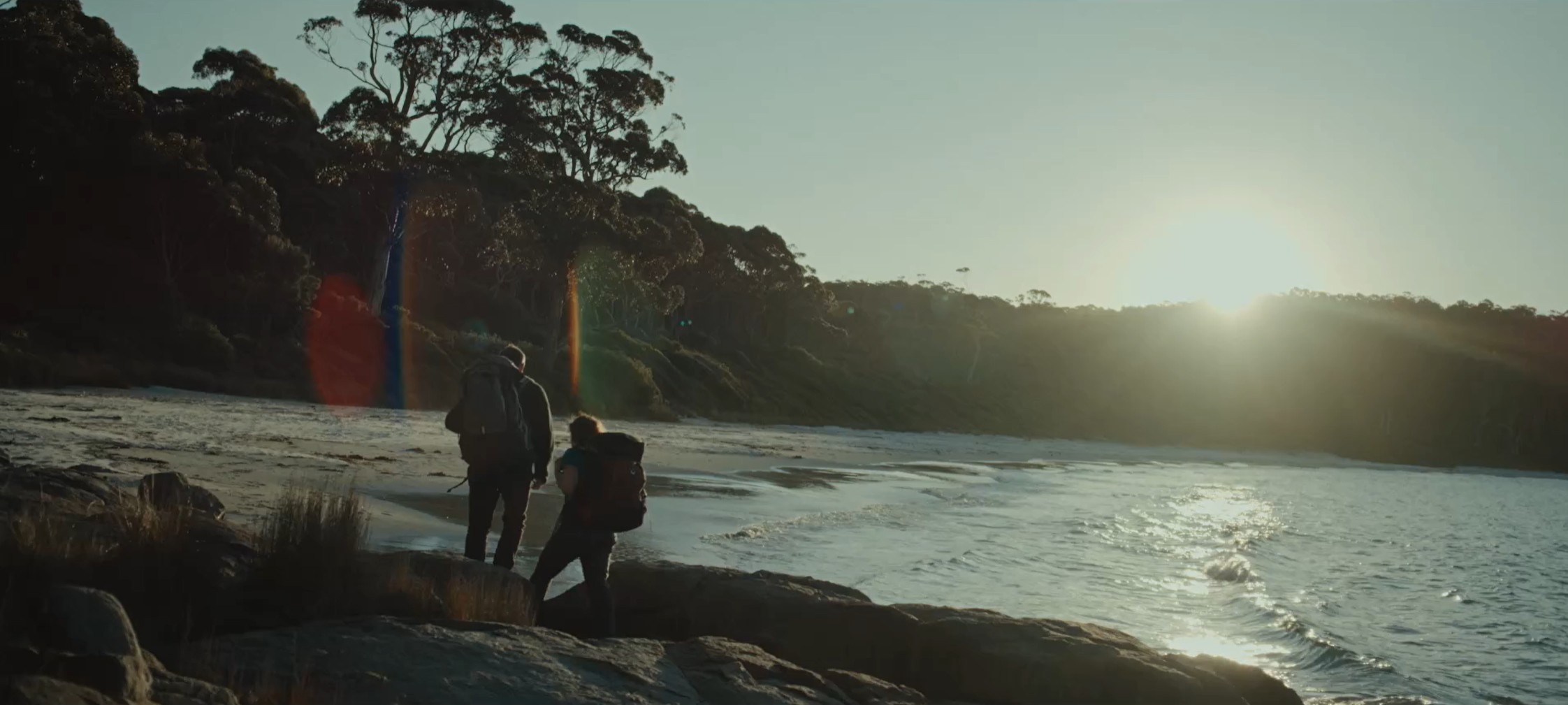 Two walkers with backpacks on rocks beside a beach in golden light, Tasmania