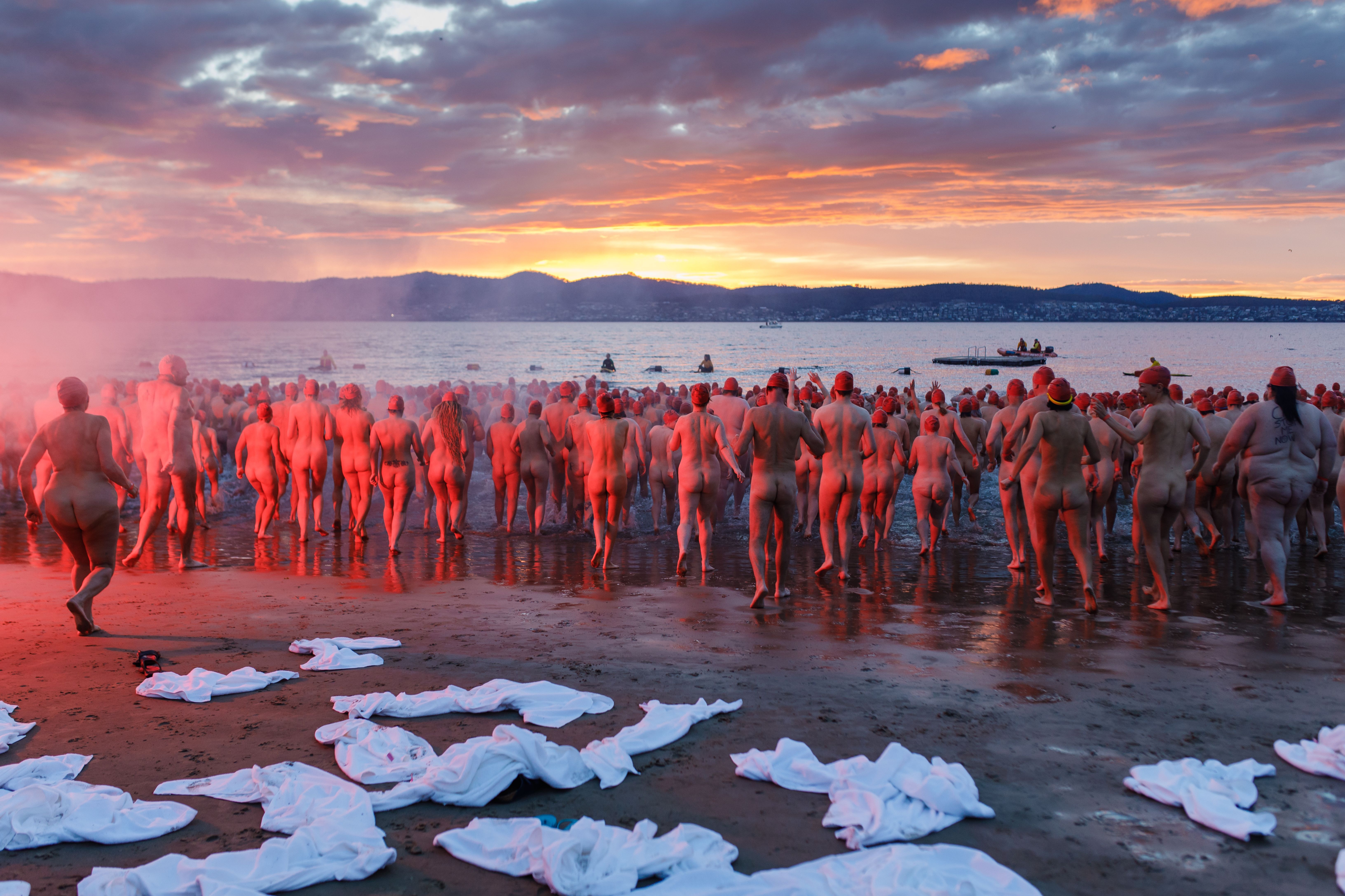 Swimmers running into the water in red caps at the Nude Solstice Swim during Dark Mofo, Long Beach, Sandy Bay, Tasmania