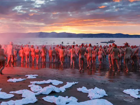Swimmers running into the water in red caps at the Nude Solstice Swim during Dark Mofo, Long Beach, Sandy Bay, Tasmania