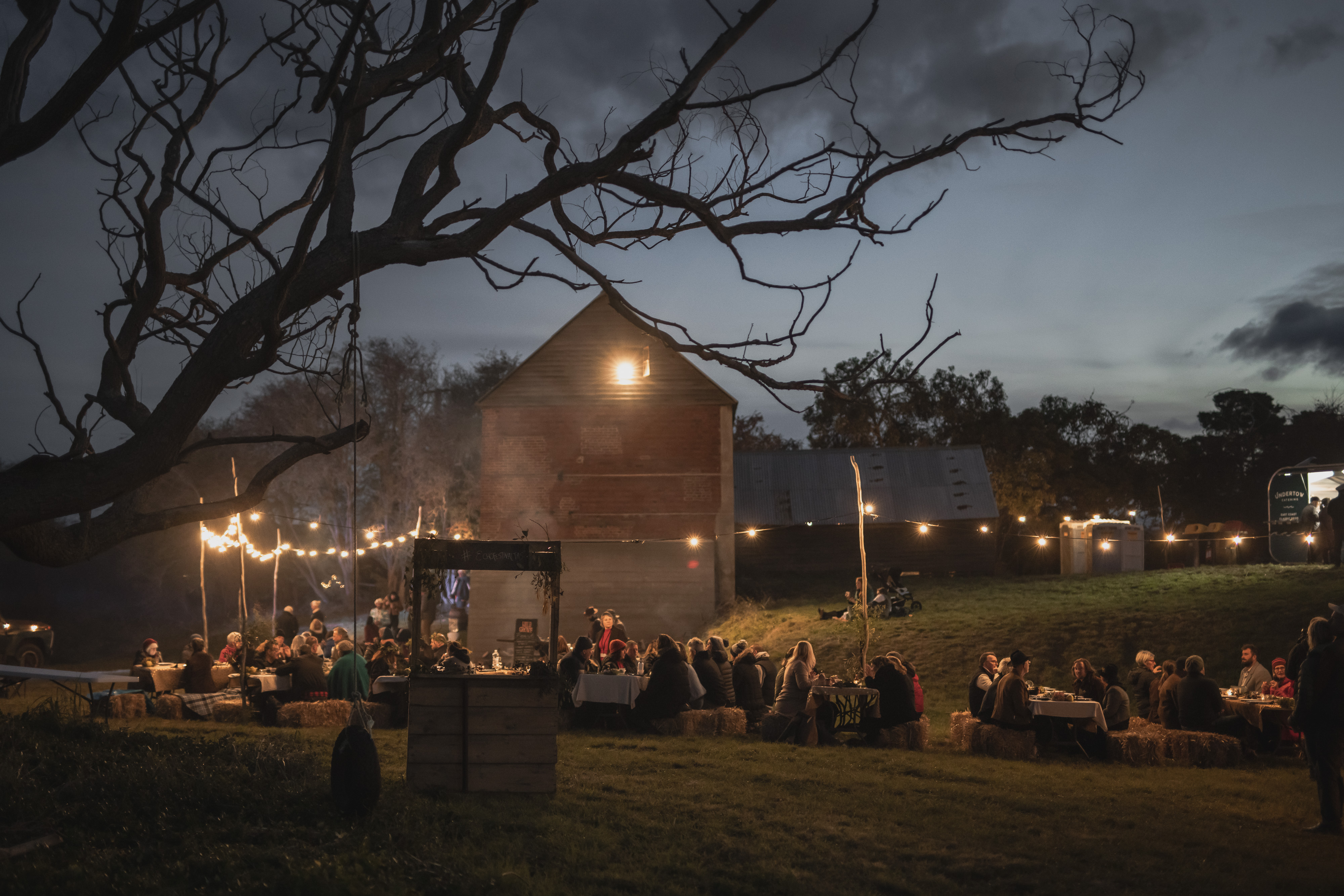 Guests at long tables under string lights at ECHO East Coast Harvest Odyssey festival at dusk, Cranbrook, Tasmania