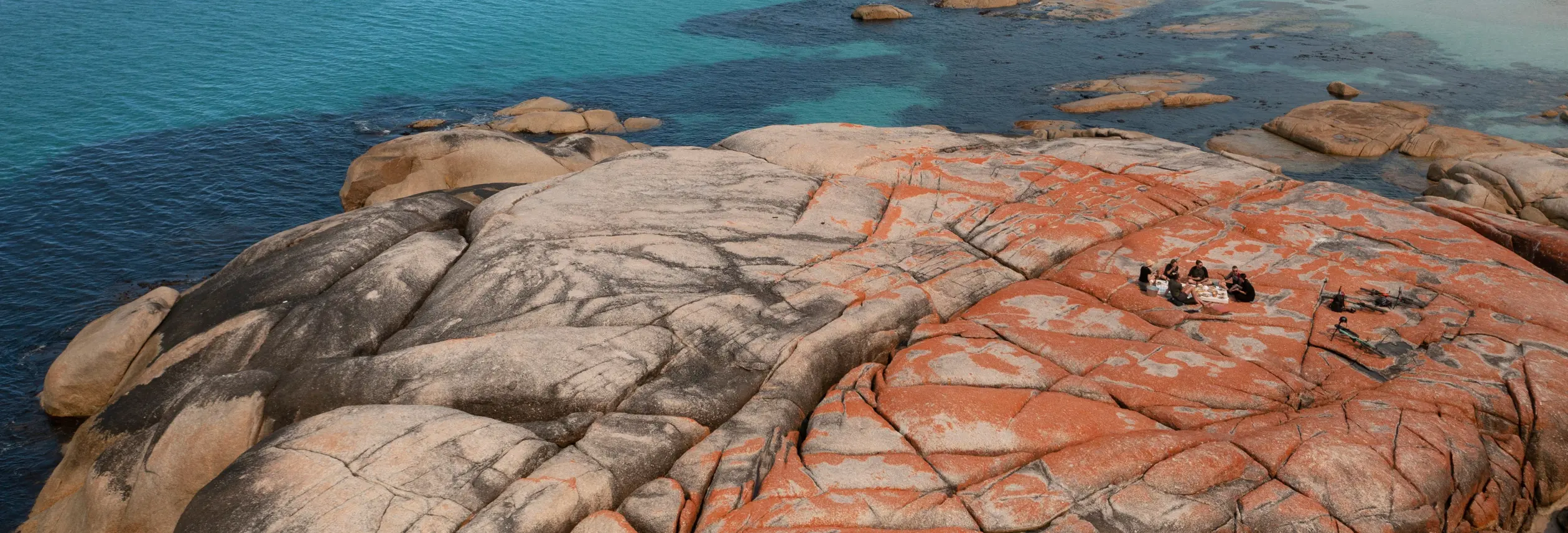 A group on lichen covered granite boulders overlooking a beach with Blue Derby Pods Ride, larapuna / Bay of Fires area