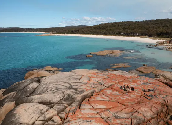 A group on lichen covered granite boulders overlooking a beach with Blue Derby Pods Ride, larapuna / Bay of Fires area