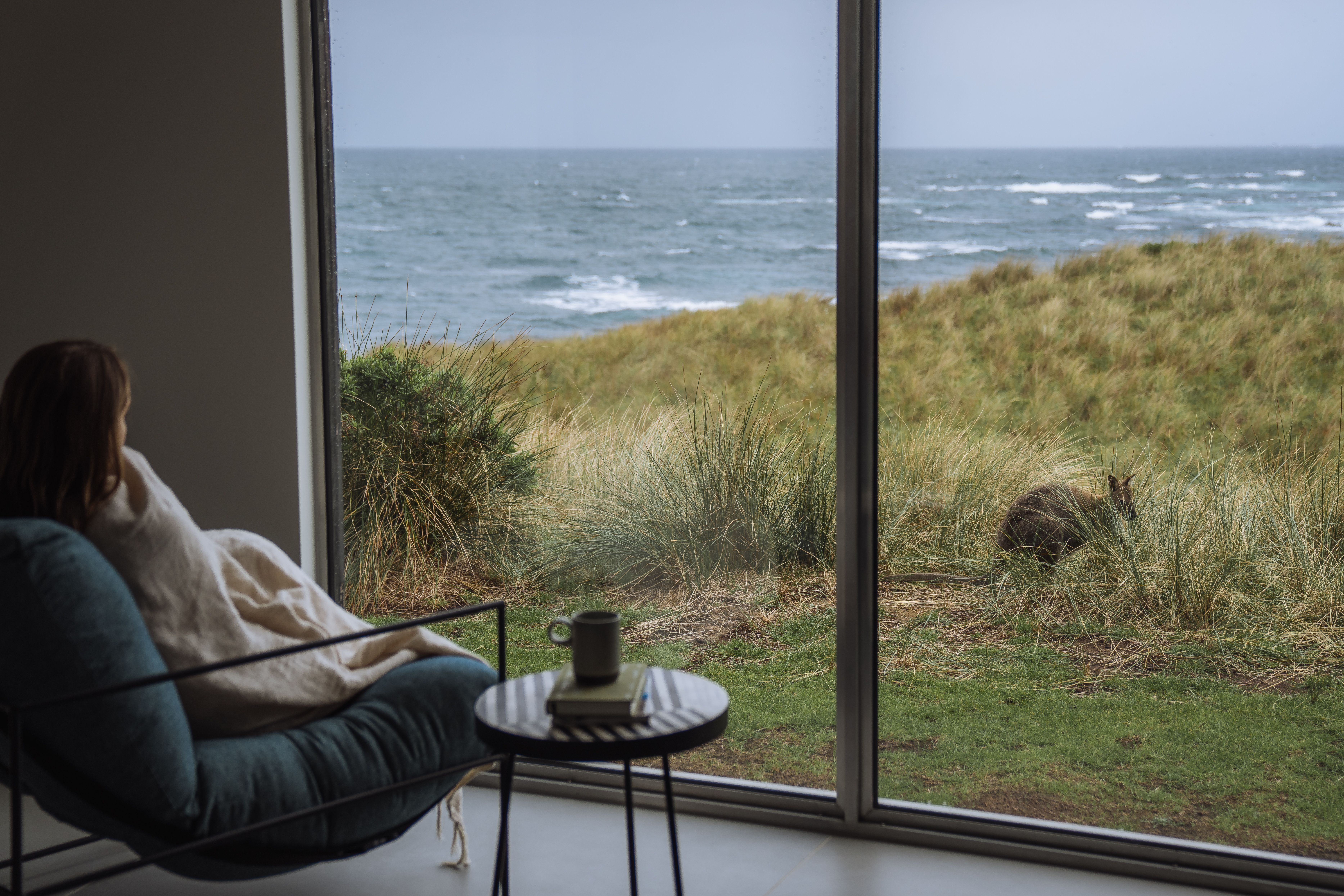 A guest wrapped in a blanket watches a wallaby in the dunes through floor-to-ceiling windows at Kittawa Lodge, King Island