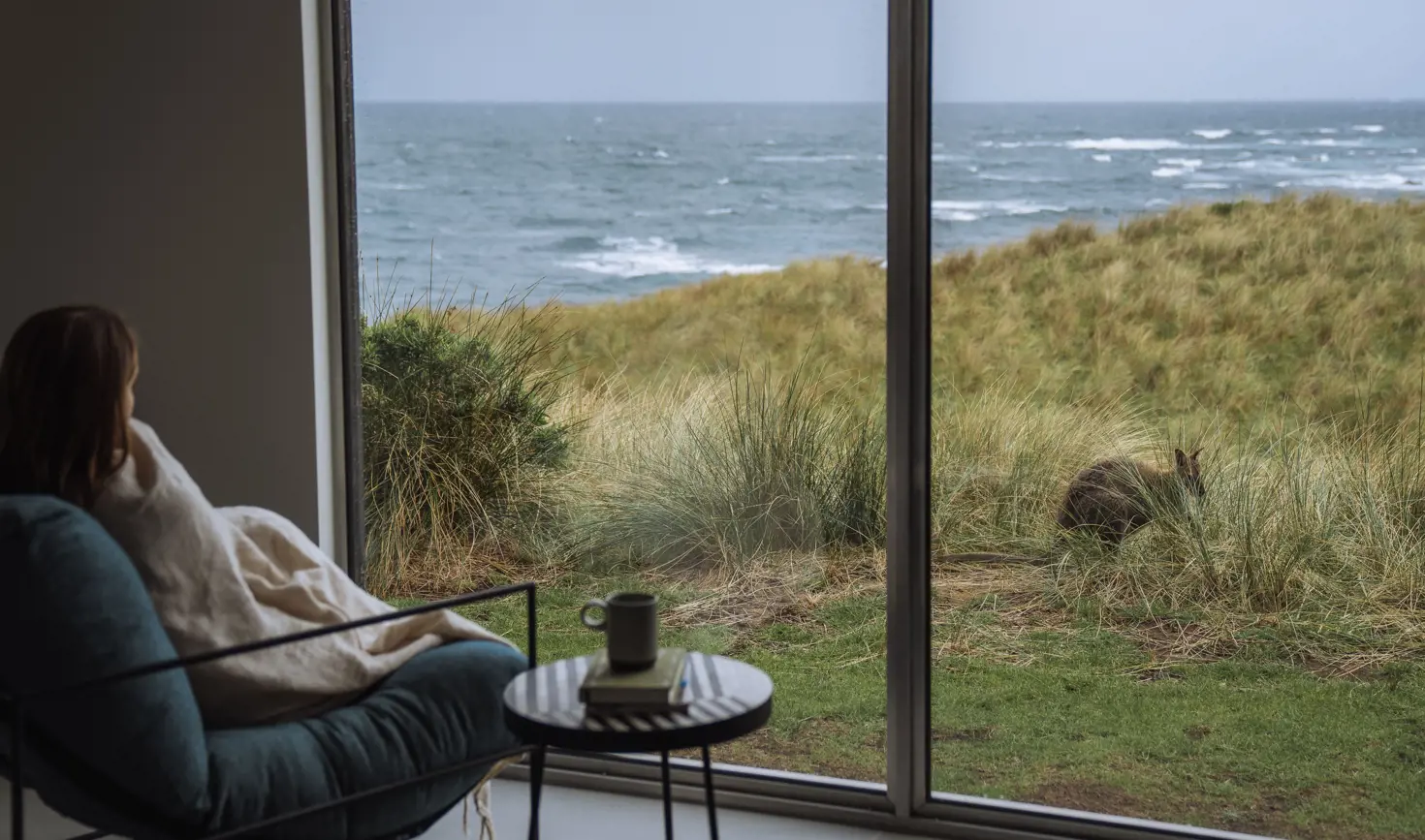 A guest wrapped in a blanket watches a wallaby in the dunes through floor-to-ceiling windows at Kittawa Lodge, King Island