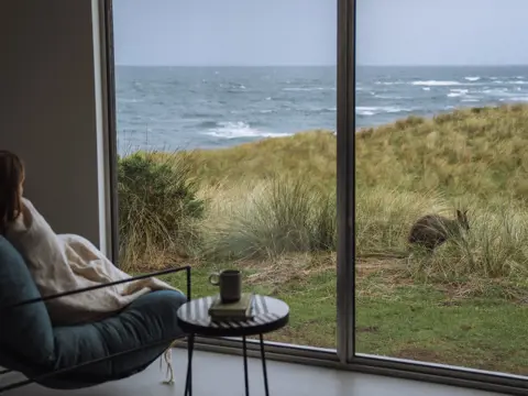 A guest wrapped in a blanket watches a wallaby in the dunes through floor-to-ceiling windows at Kittawa Lodge, King Island