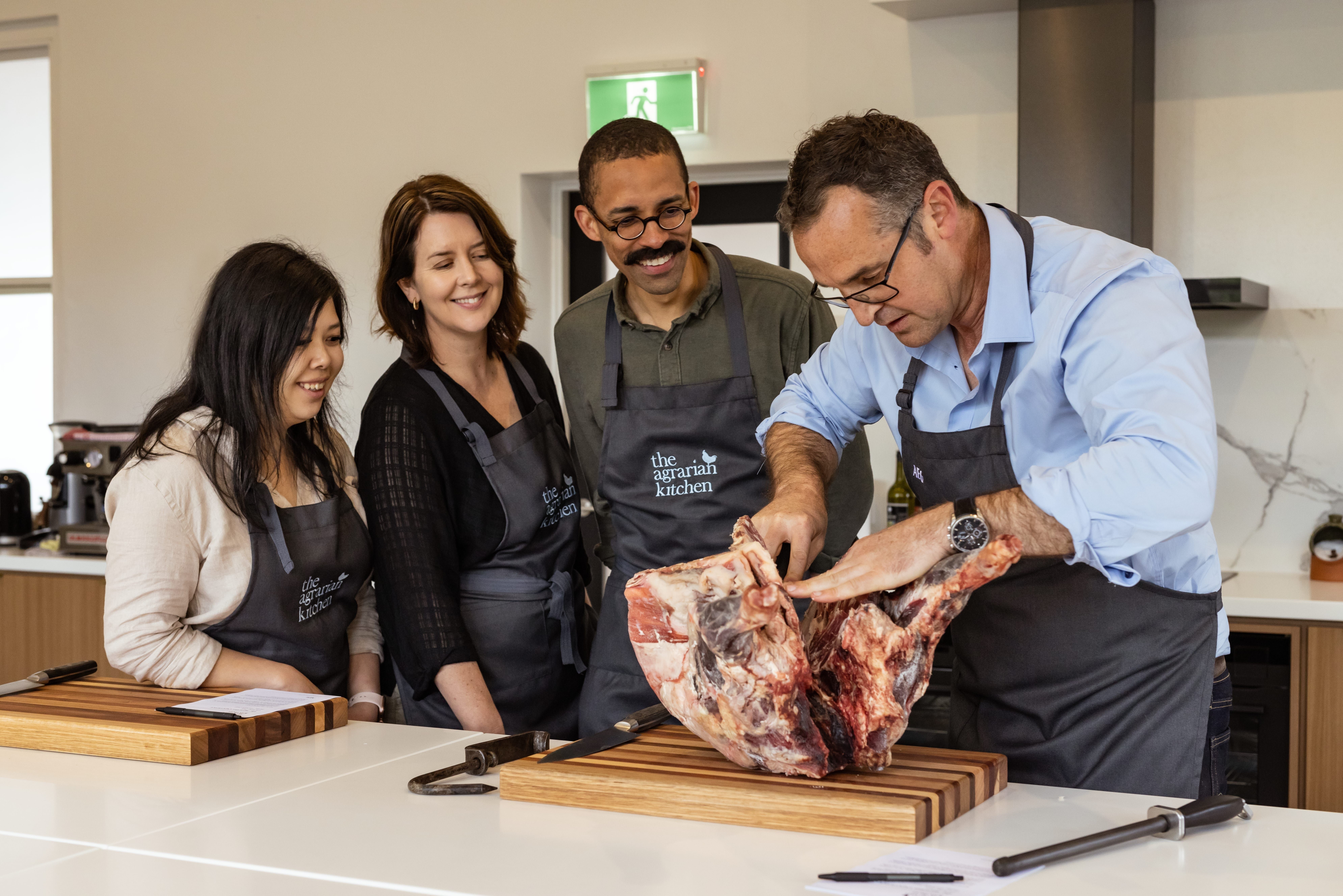 A chef demonstrating butchery to three students in aprons at The Agrarian Kitchen cooking class, New Norfolk