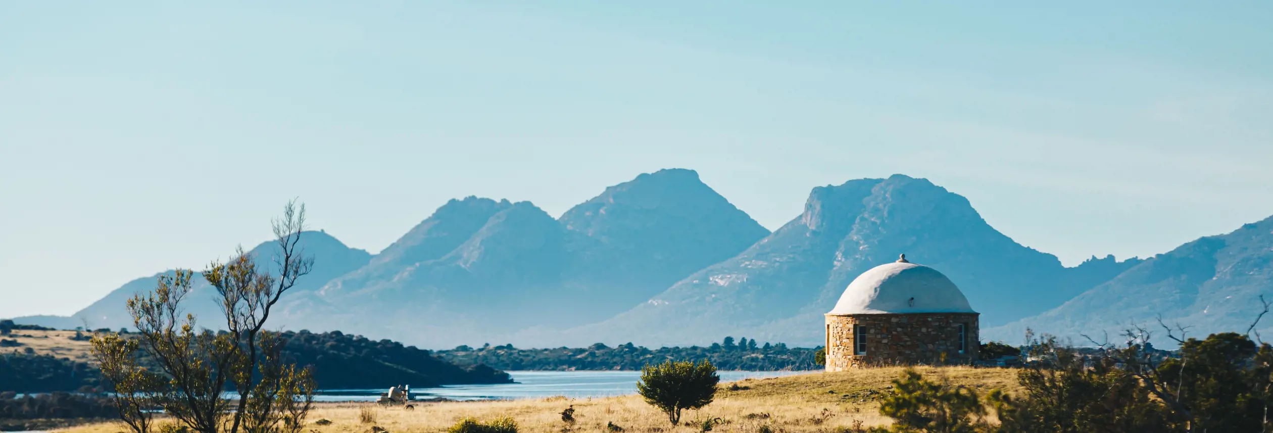 A round stone building with a domed roof in open grassland with water and mountains beyond at The Bend Vineyard, Swansea