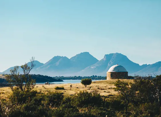 A round stone building with a domed roof in open grassland with water and mountains beyond at The Bend Vineyard, Swansea