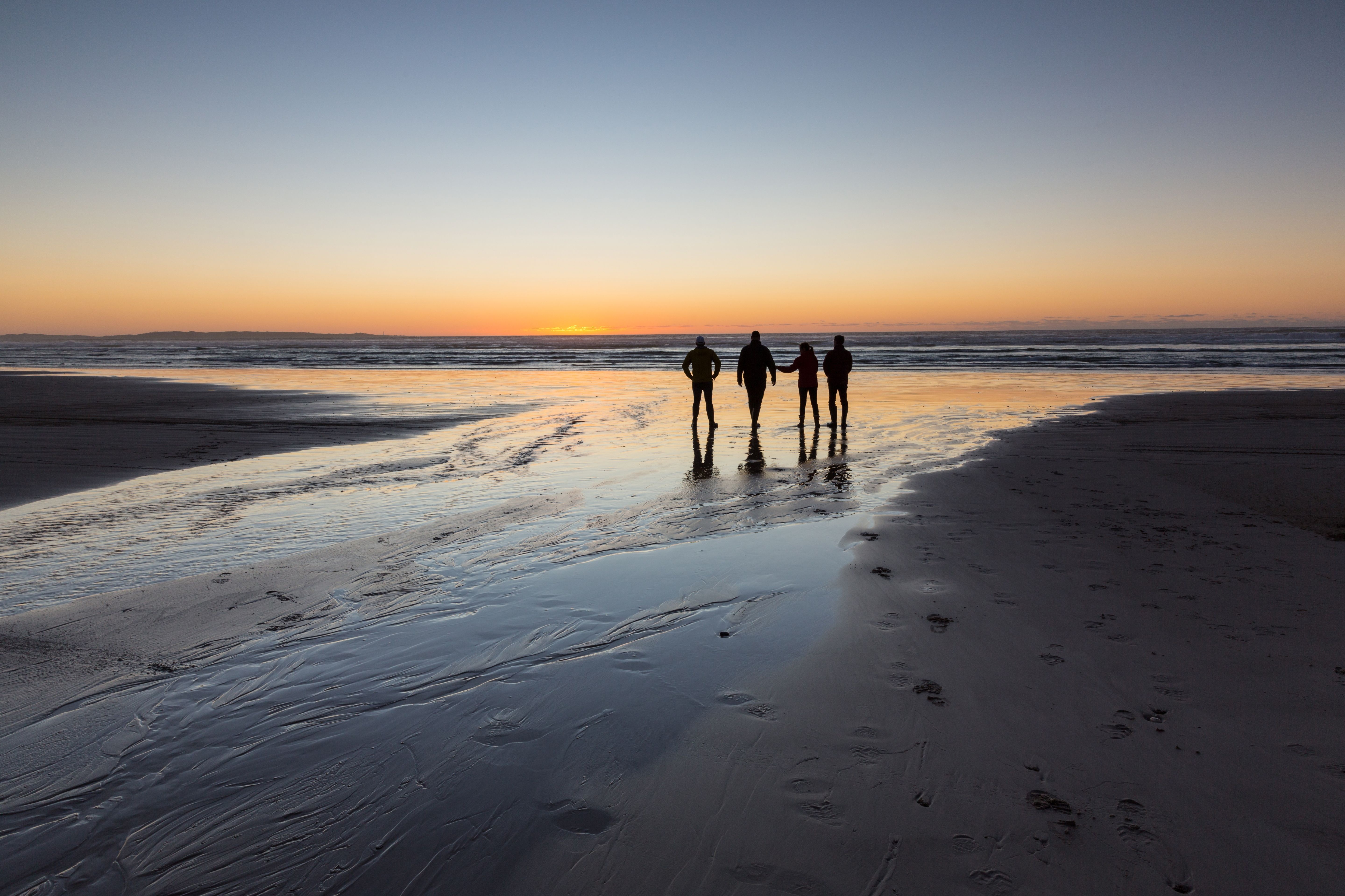 Four people silhouetted on Ocean Beach watching the sun set over the water, Strahan