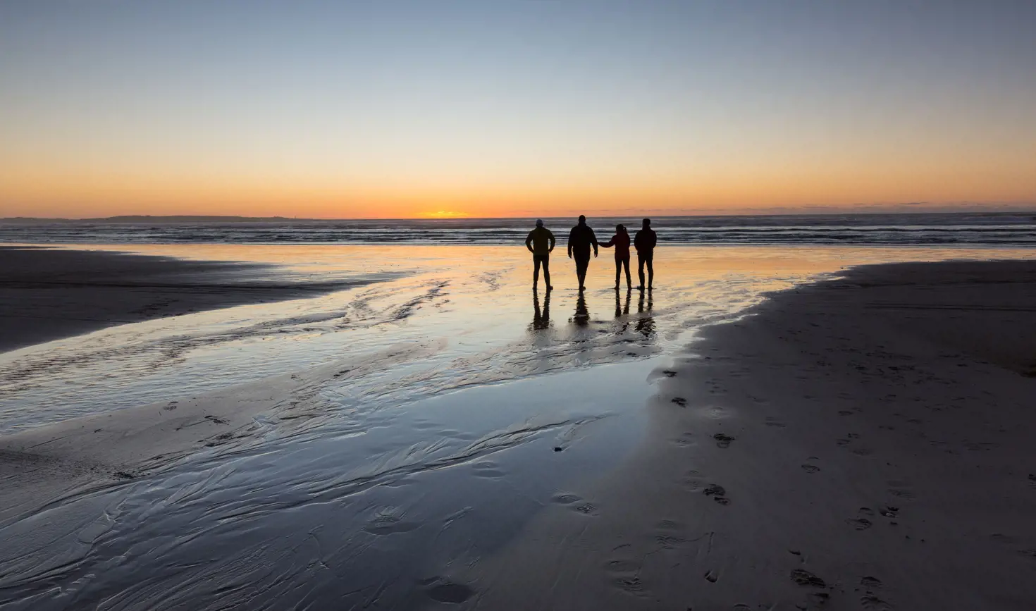Four people silhouetted on Ocean Beach watching the sun set over the water, Strahan