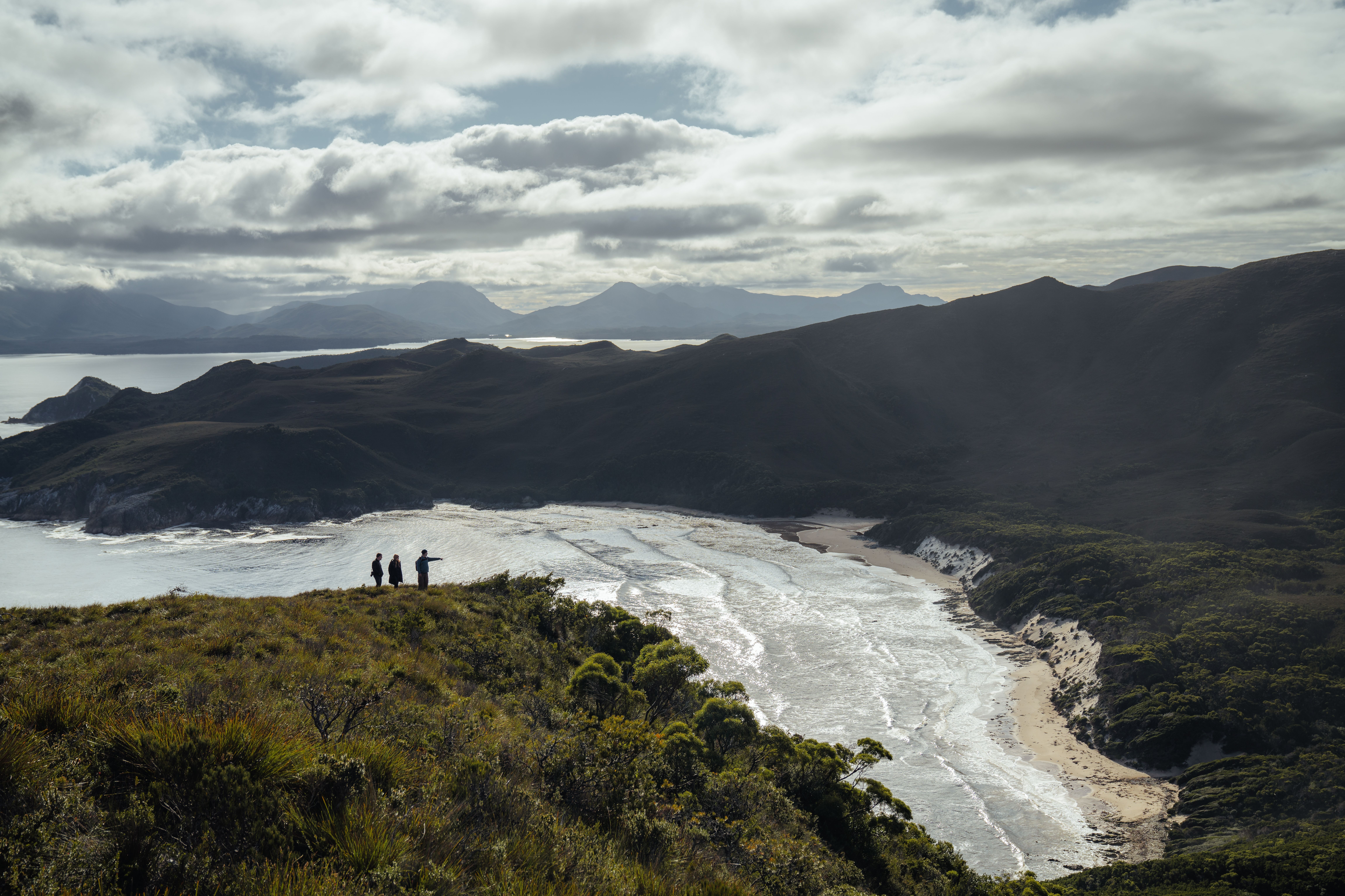 Three walkers on a clifftop overlooking a sweeping beach and mountain ranges in Southwest National Park