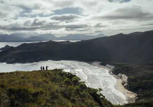 Three walkers on a clifftop overlooking a sweeping beach and mountain ranges in Southwest National Park