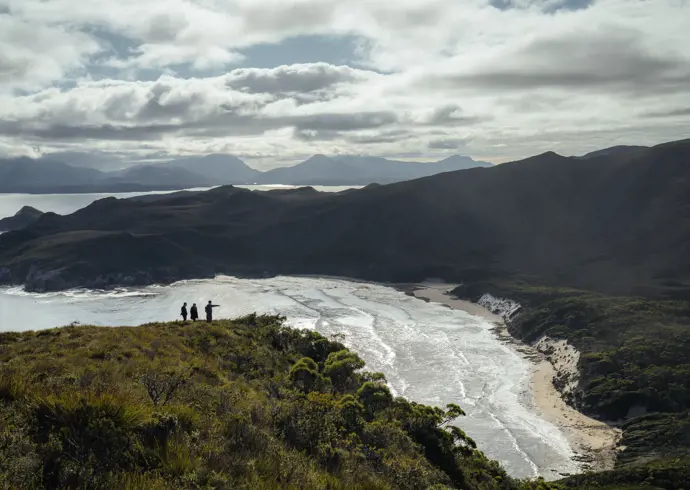 Three walkers on a clifftop overlooking a sweeping beach and mountain ranges in Southwest National Park