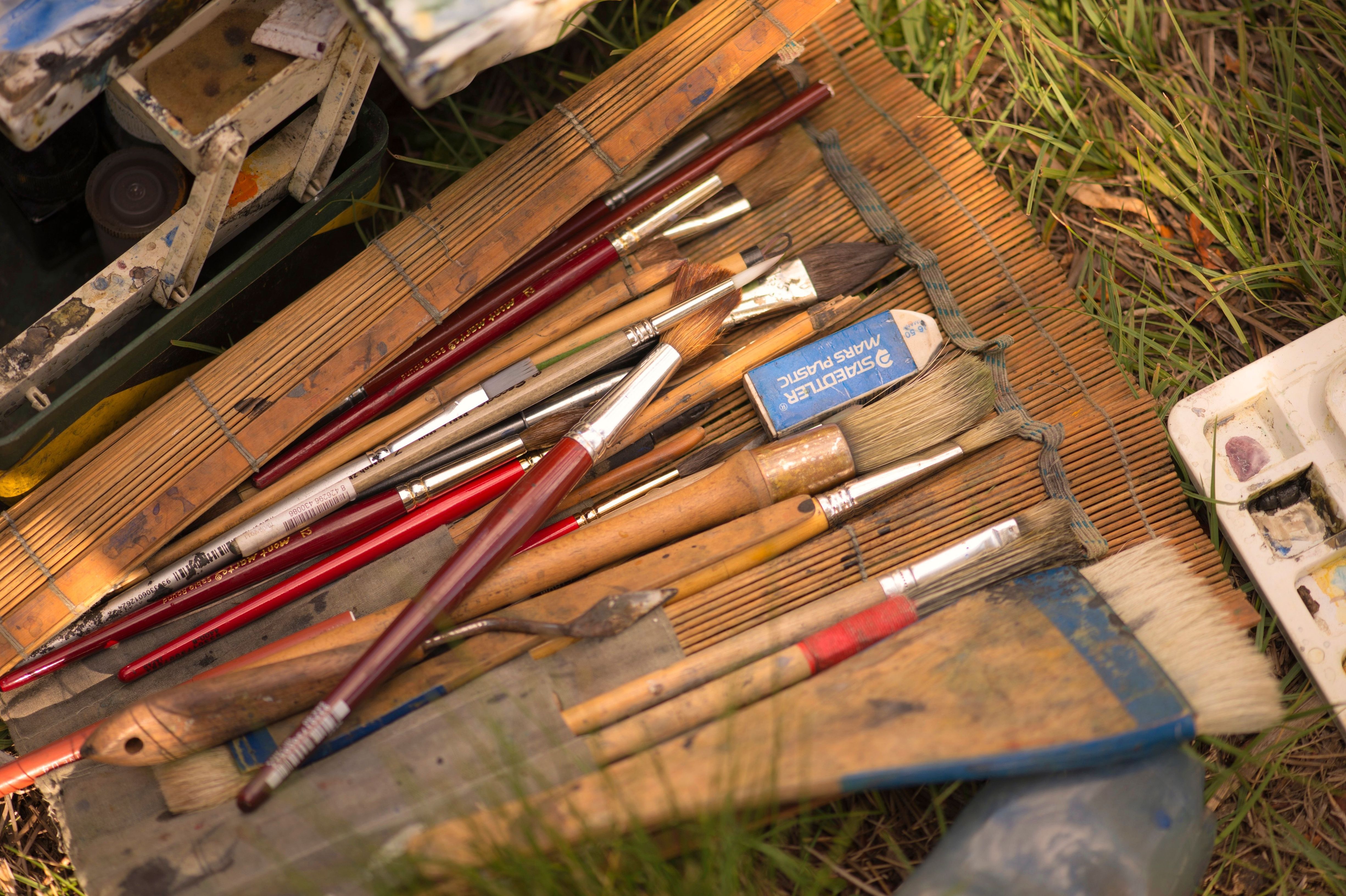 An artist's paintbrushes and palette laid out on a bamboo mat in the grass