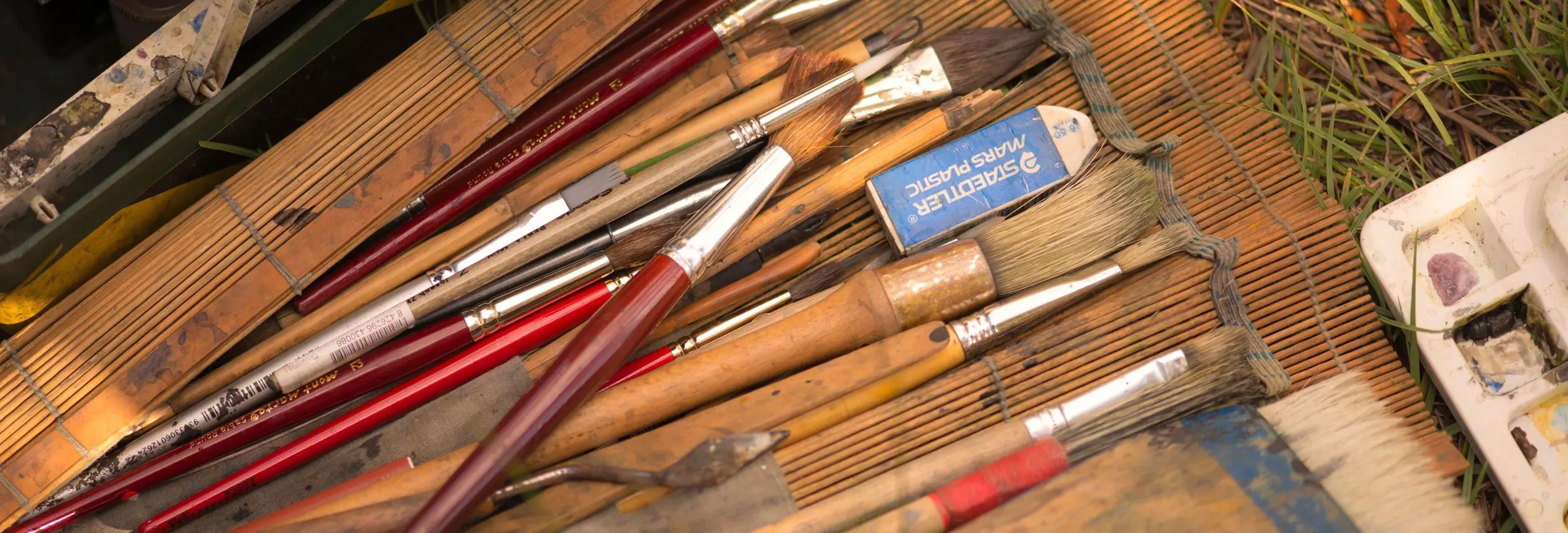 An artist's paintbrushes and palette laid out on a bamboo mat in the grass